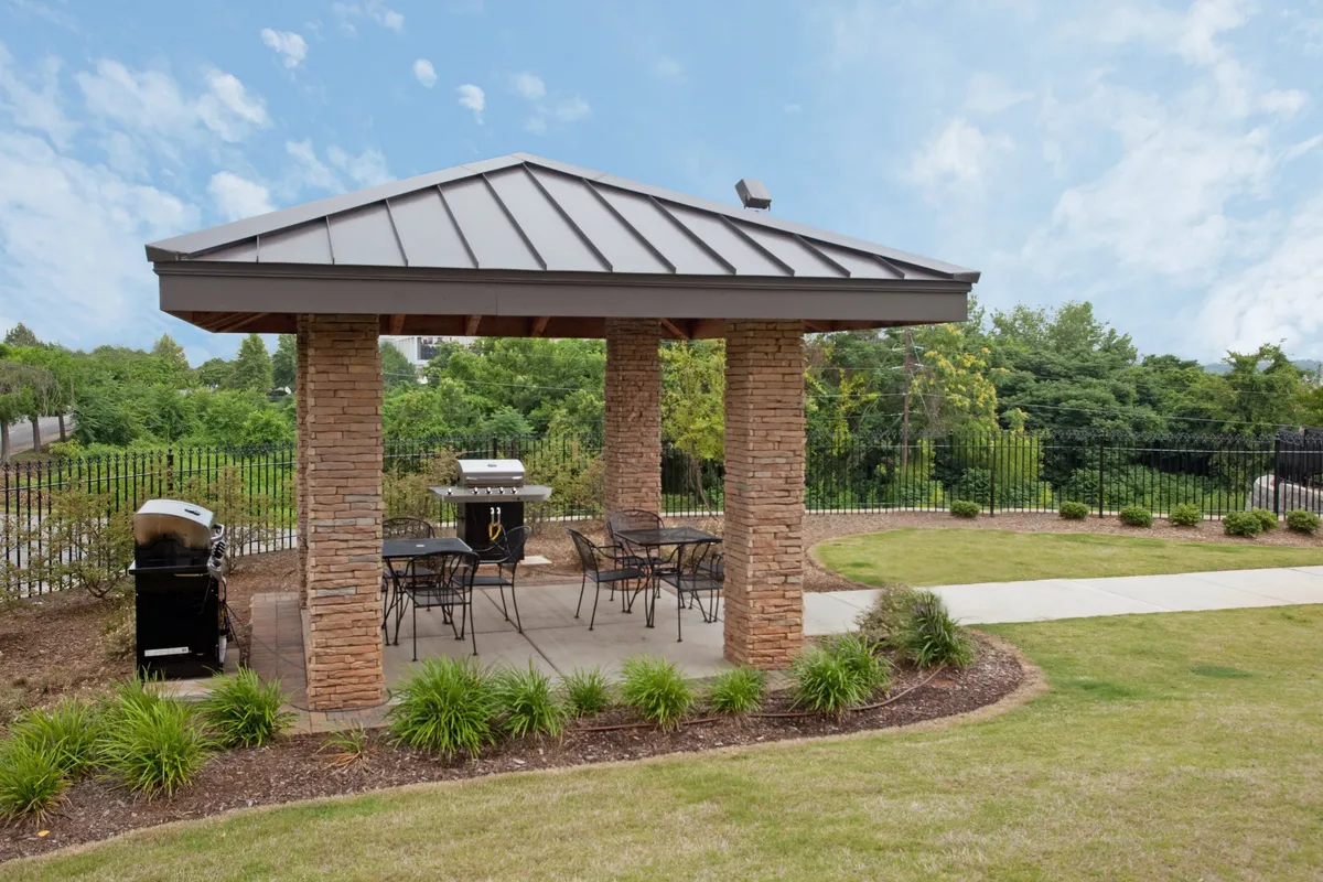 This image depicts an inviting exterior communal space with a grill, comfortable seating, stone columns, and landscaped greenery. The atmosphere is relaxed and communal, ideal for social gatherings, barbecues, and outdoor dining. The sunset lighting further enhances the welcoming vibe.