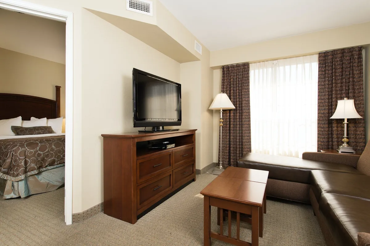 The image features a cozy, well-lit living area inside a modern condo suite. A plush sofa is arranged facing a large flat-screen television, flanked by elegant lamps and soft curtains, creating an inviting space ideal for relaxing after a day exploring downtown Columbia. The atmosphere is warm and comfortable, suggesting a tranquil retreat for couples or solo travelers.
