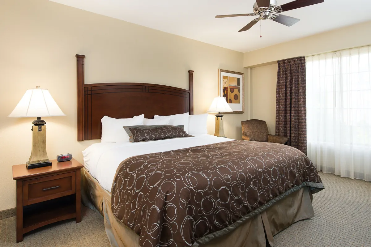 This image showcases a spacious king-size bed adorned with decorative pillows and flanked by elegant table lamps. Natural light filters into the room, illuminating the cozy bedding and stylish, individualized touches that hint at the suite's unique décor.