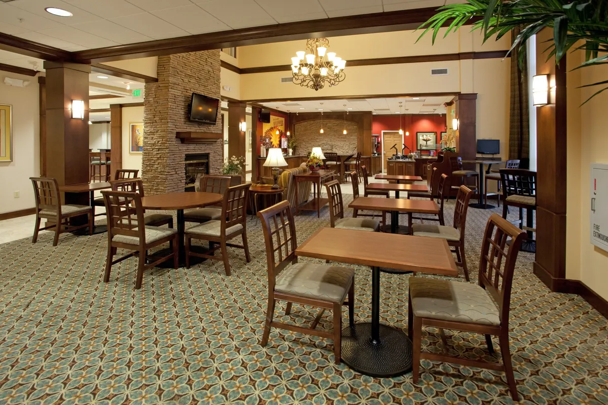 This inviting living area features a warm fireplace, wooden tables, a seating arrangement with comfortable chairs, and a television mounted above the hearth. The scene suggests a space ideal for both socializing and relaxing, with ample room to read, dine, or enjoy entertainment.