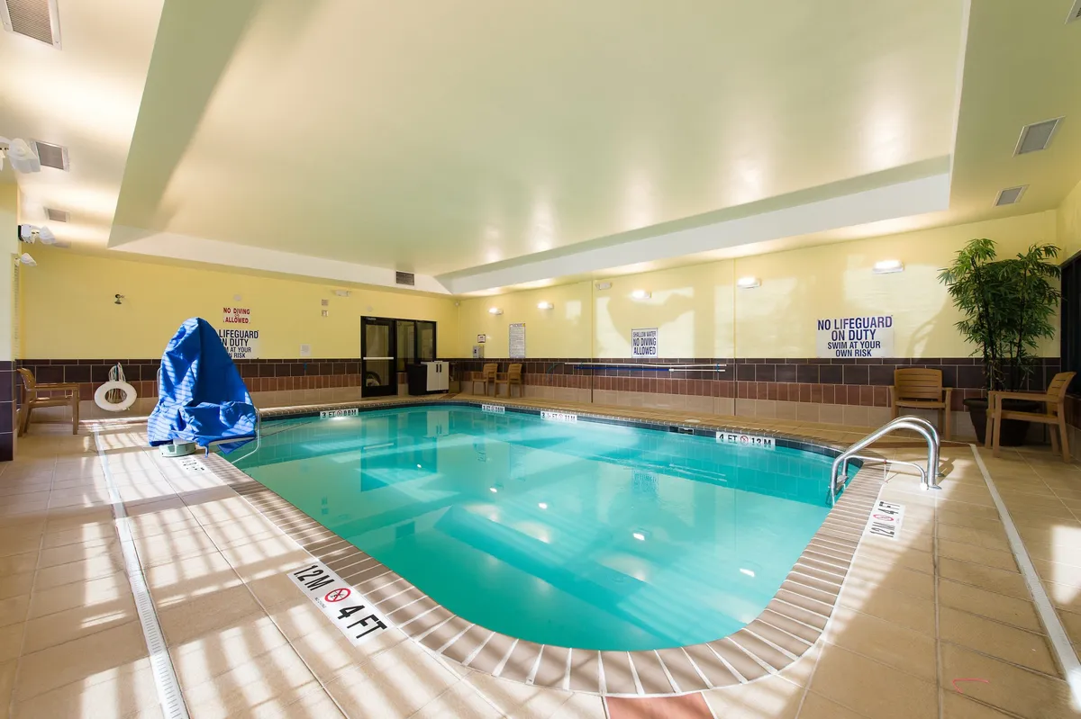 The image features a heated indoor swimming pool with a blue poolside cover, lounge chairs, and safety signage. There are children playing in the water and adults lounging nearby, creating a refreshing and relaxed mood. Evening lighting gives the space a cozy, welcoming ambiance.