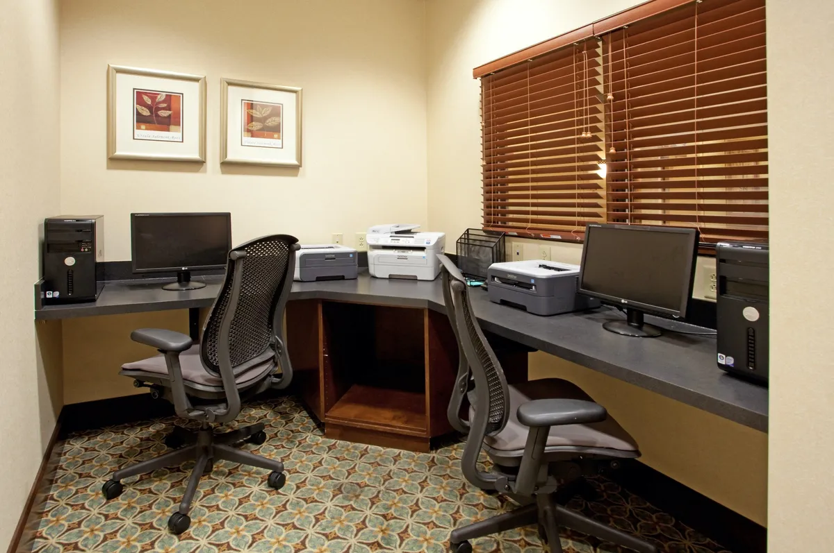 A modern business center equipped with several computers, printers, office chairs, and tasteful artwork. The scene conveys an atmosphere of productivity and professionalism, with guests using the technology for work or collaboration.