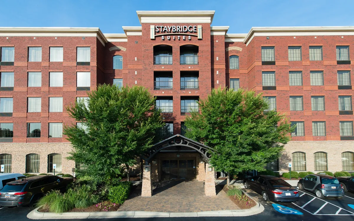 This exterior image showcases the inviting entrance awning of Staybridge Suites Columbia, set against a backdrop of large windows and brick facade, surrounded by landscaped greenery. Guests can be seen arriving, hinting at easy access and the welcoming nature of the property.