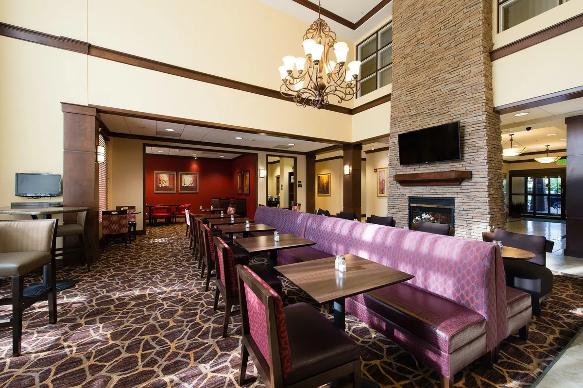 A tastefully decorated lounge area with a chandelier, a warming fireplace, and a thoughtfully arranged seating area. Decorative artwork adorns the walls, and guests are pictured engaging in social and individual activities—some chatting, others working or reading.