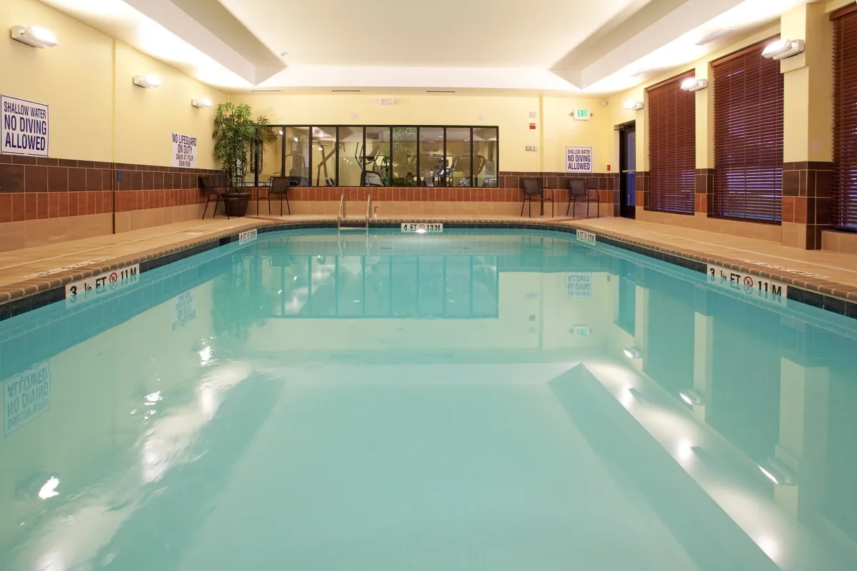 This image features a heated indoor pool as the central focus, with poolside seating for relaxation and a glimpse of the fitness area visible in the background. The tranquil mood captures both guests swimming and others unwinding by the water, while a few patrons can be seen working out behind the pool area.
