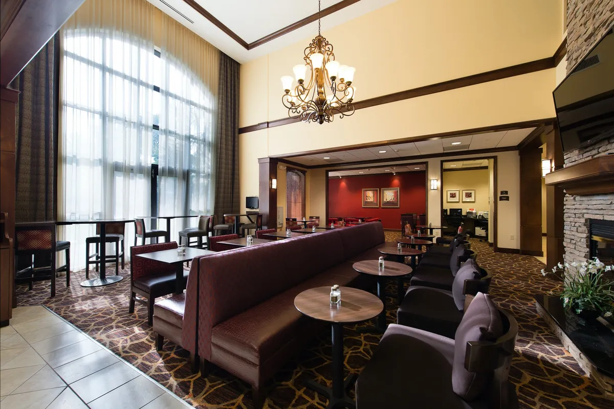 A spacious, welcoming lobby area featuring a large chandelier, floor-to-ceiling windows that allow natural light to flood the space, comfortable seating, and a central fireplace. The environment is cozy yet open to the outside world, suggesting a communal hub where guests gather before and after exploring the city.