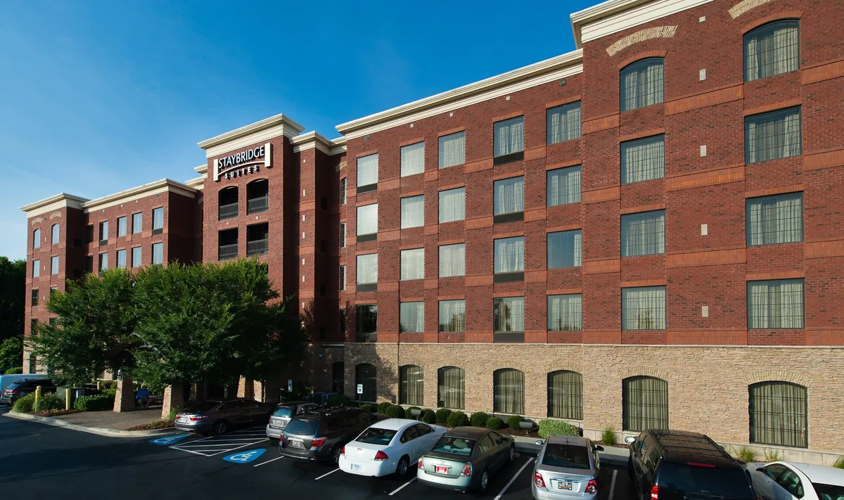 An exterior shot highlighting the building facade, entrance sign, and adjacent parking area. The visual narrative includes guests checking in, unpacking luggage, and hints at the property's strategic location for exploring downtown and accessing corporate venues.