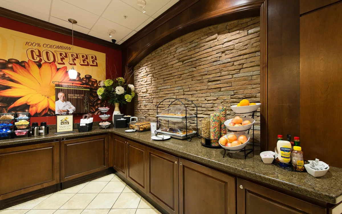 A cozy coffee station with a display of baked goods, fresh fruit, and a decorative floral arrangement set against a stylish stone wall. The scene exudes warmth and hospitality, emphasizing the availability of morning treats and coffee.